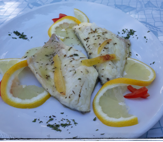 Filetti di pesce bianco grigliati con fette di limone e prezzemolo su un piatto bianco.
