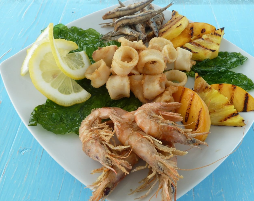 Piatto di frutti di mare con calamari, pesce fritto, fette grigliate, spicchi di limone e prezzemolo su un piatto bianco.
