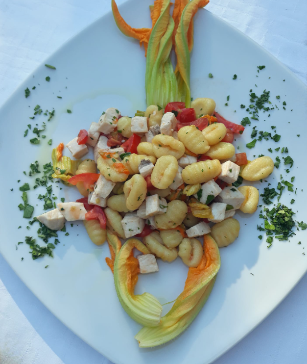 Gnocchi impiattati con verdure a dadini ed erbe aromatiche, guarniti con petali di fiori su un piatto quadrato bianco.