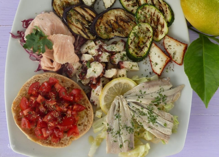 Piatto di mezze greche con hummus, tzatziki, melanzane, zucchine, pomodori e spicchi di limone.