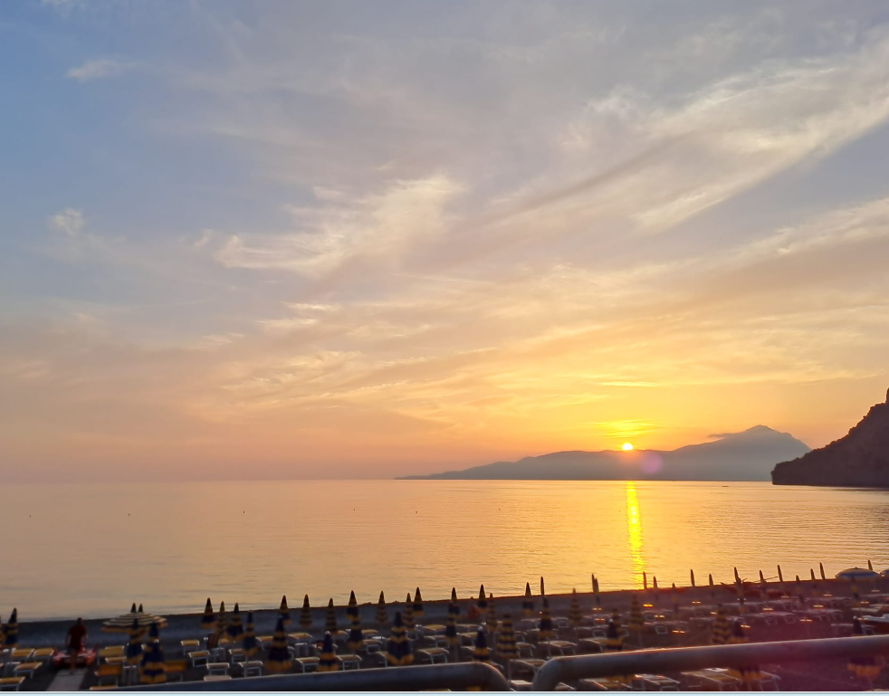 Tramonto su un mare calmo con lettini e ombrelloni sulla spiaggia, riflessi dorati sull'acqua.