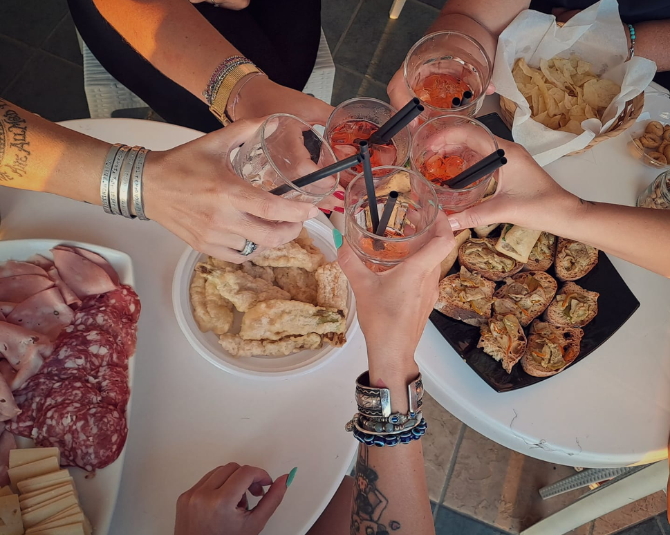 Mani che alzano cocktail attorno a un tavolo con stuzzichini, tra cui carne, pane e bocconcini ripieni.