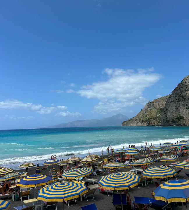 Spiaggia soleggiata con ombrelloni a righe, onde turchesi e scogliere rocciose sotto un cielo azzurro.