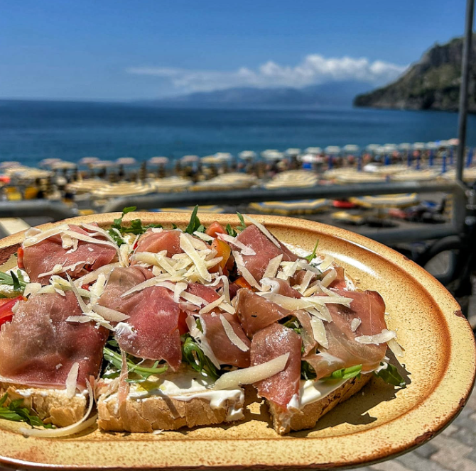 Piatto di bruschette con prosciutto crudo su un tavolo in riva al mare, con l'oceano e le scogliere sullo sfondo.