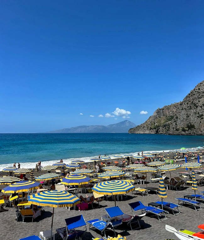 Spiaggia soleggiata con ombrelloni a righe e lettini, affacciata su un mare turchese e una costa rocciosa.
