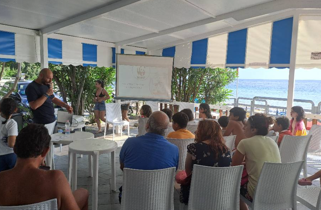 Persone sedute in una tenda a strisce assistono a una presentazione con schermo di proiezione e altoparlante.