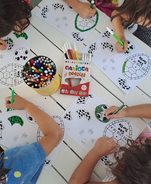 Dei bambini colorano dei fogli di lavoro attorno a un tavolo con pennarelli, pastelli e una scatola di matite Color'Peps.