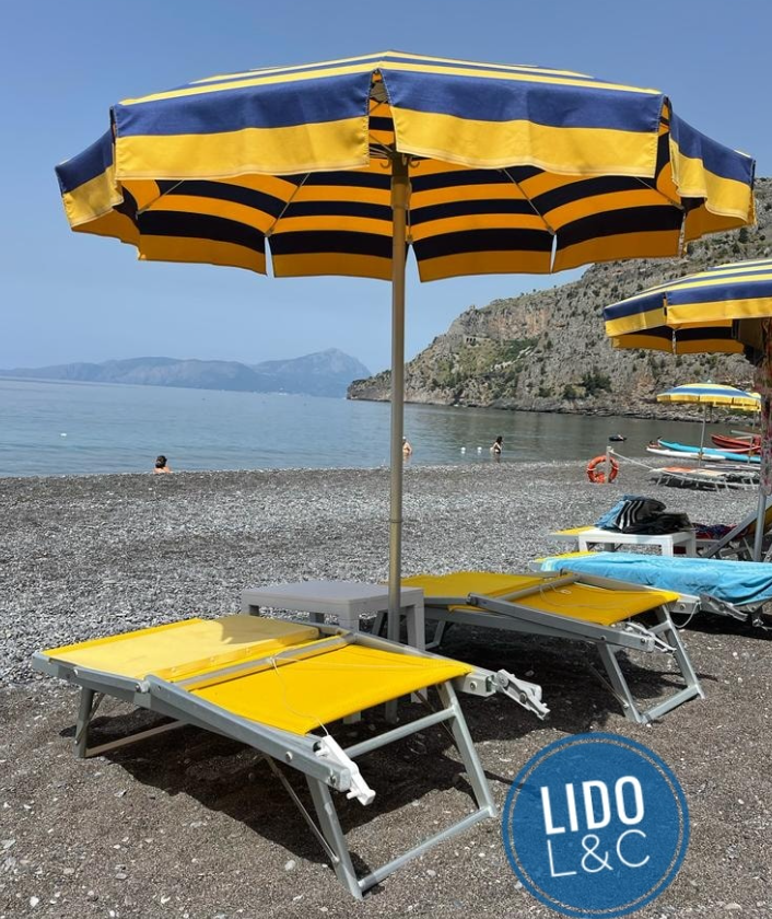 Lettini e ombrellone a strisce gialle su una spiaggia di ciottoli, con acque tranquille e montagne sullo sfondo.
