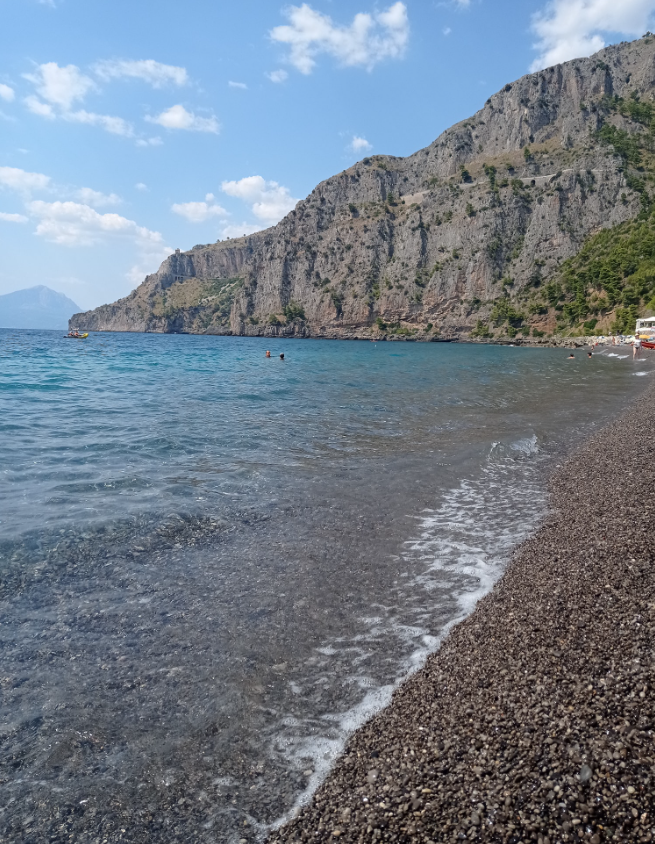 Spiaggia rocciosa con acqua cristallina, riva di ciottoli e scogliere a picco sotto un cielo parzialmente nuvoloso.