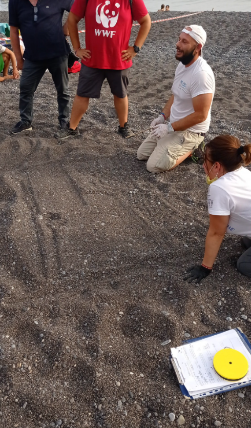 Persone inginocchiate su una superficie di ghiaia durante un'attività in cantiere, con un blocco appunti e un disco giallo in primo piano.