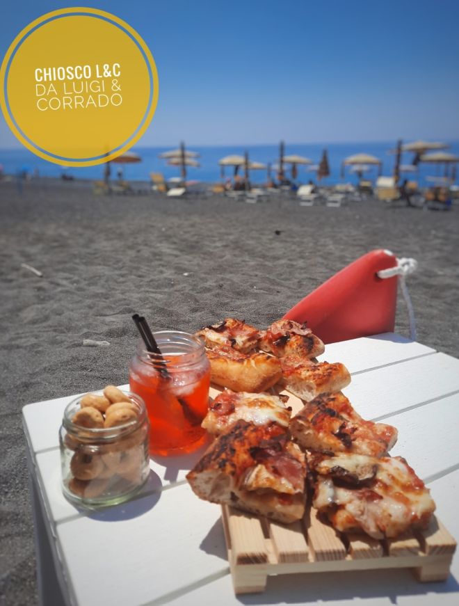 Pizza e bevande in riva al mare, servite su un tavolo con il mare e gli ombrelloni sullo sfondo.
