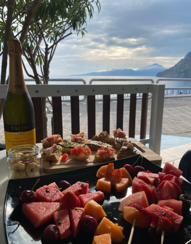 Pranzo all'aperto in riva al mare con piatti di frutta e una bottiglia di vino su un tavolo con vista sull'acqua.