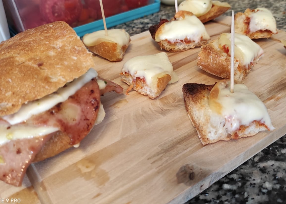 Panini e pasticcini salati monoporzione ricoperti di formaggio fuso, serviti su un tagliere di legno.