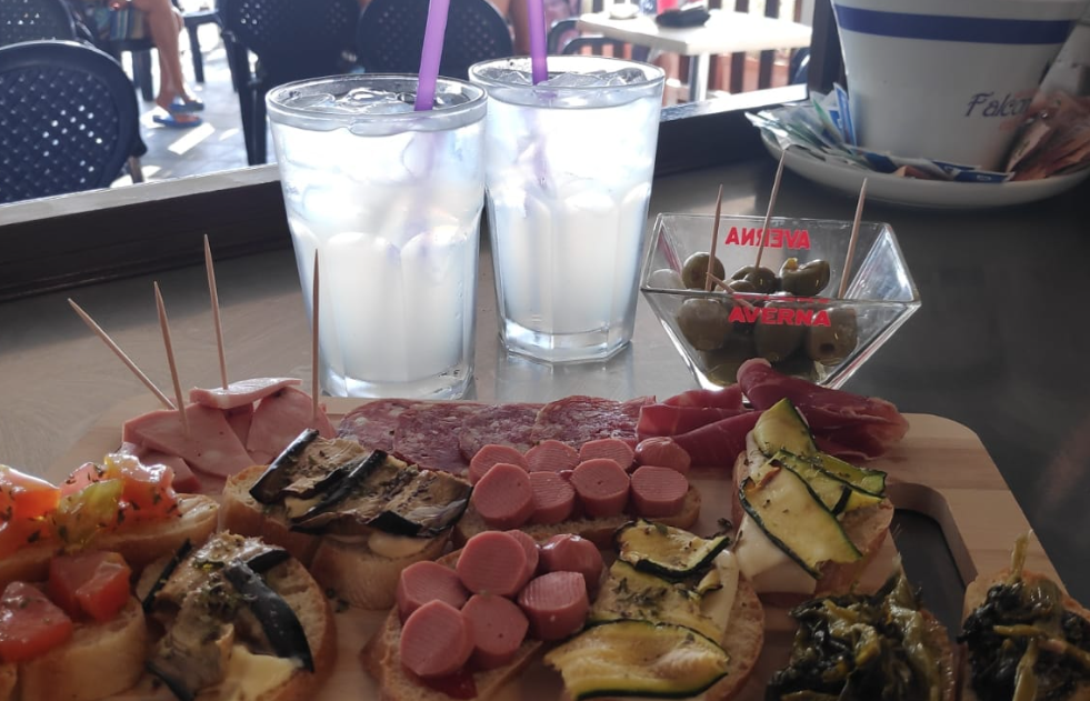 Tagliere di salumi con affettati, formaggio e zucchine su un tavolino da caffè, con due bevande ghiacciate e una ciotola sullo sfondo.