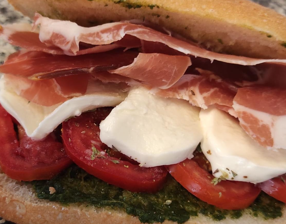 Primo piano di un panino con prosciutto, mozzarella, pomodori e pesto su una baguette.