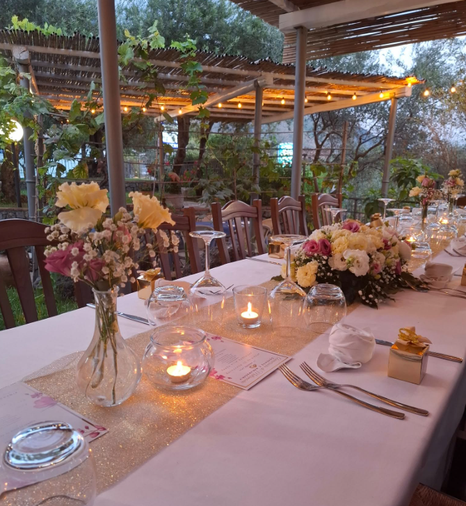 Elegante tavolo da pranzo all'aperto con tovaglia bianca, candele e centrotavola floreali in un ambiente da giardino.