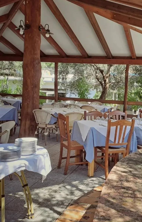Ristorante all'aperto con tavoli vuoti, sedie di legno e un tetto di paglia che si affaccia sul verde.