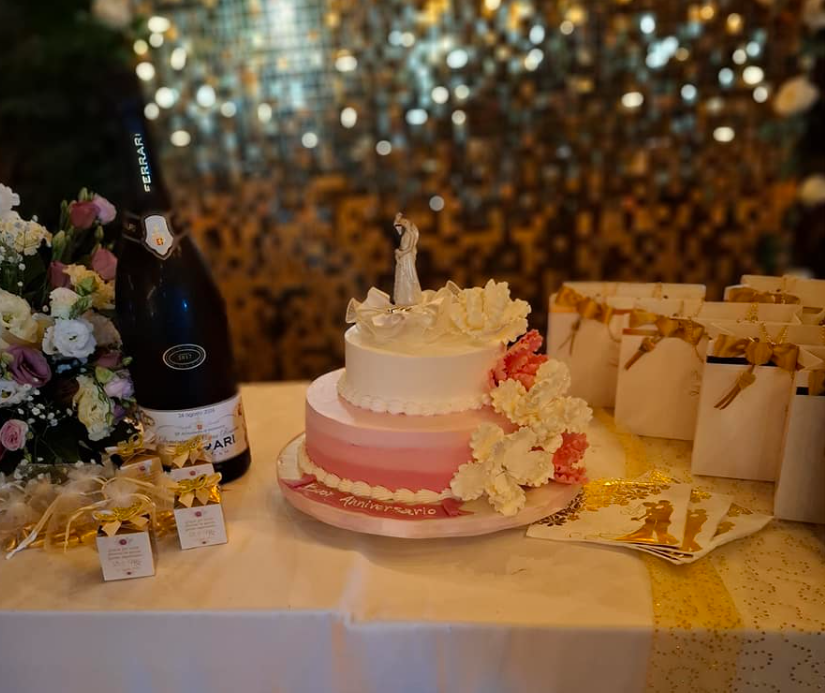 Torta a più piani rosa su un tavolo decorato con fiori, sacchetti regalo e una bottiglia di champagne durante una festa.