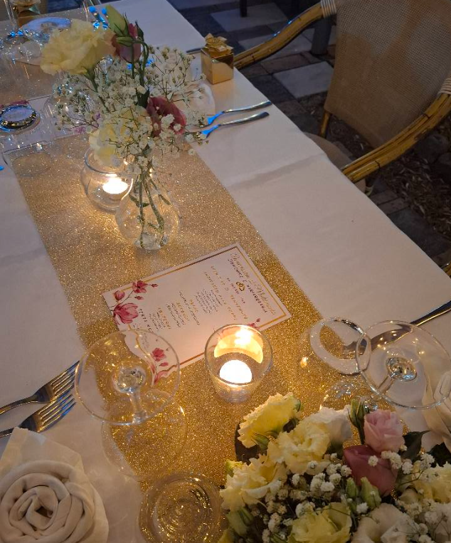 Elegante apparecchiatura della tavola con runner dorato, fiori, candele e segnaposto su una tovaglia bianca.