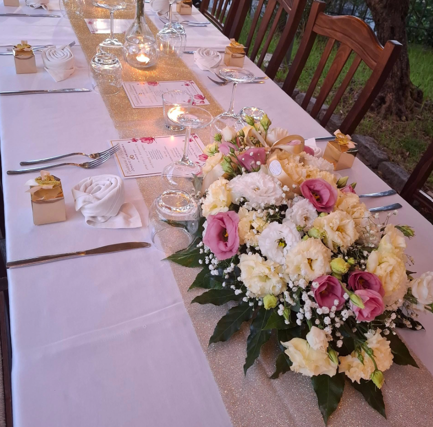 Tavolo da pranzo all'aperto decorato con tovaglie bianche, candele e centrotavola floreale rosa e bianco.