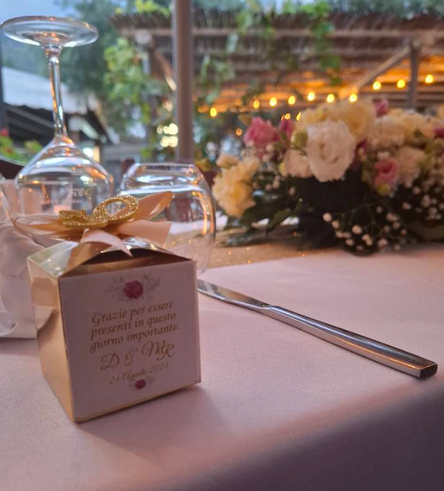 Elegante cofanetto regalo e penna d'argento su un tavolo illuminato da candele, con centrotavola floreale e calice di vino capovolto sullo sfondo.