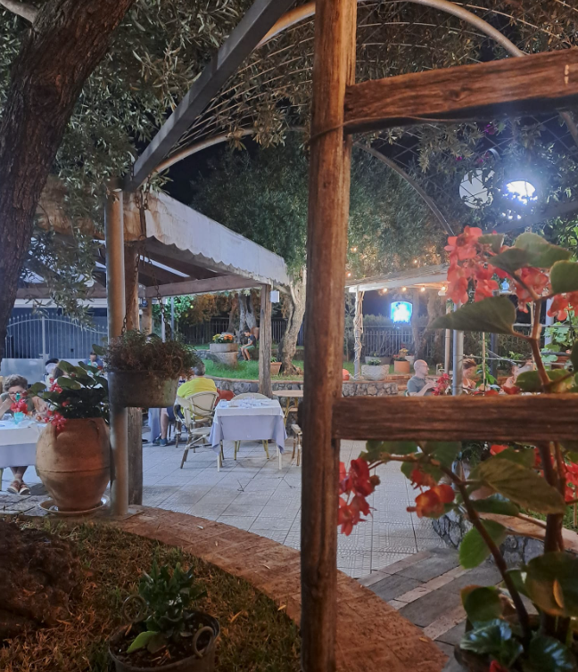 Dehor di un caffè all'aperto di sera, con tavoli e sedie bianchi, travi in legno e fiori rossi illuminati da luci calde.