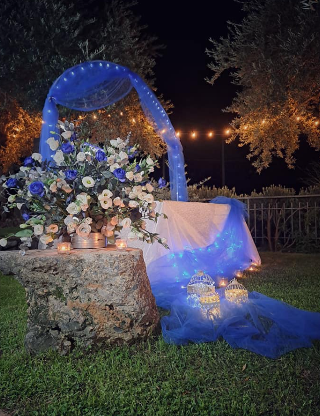 Un arco nuziale floreale drappeggiato di blu, posto accanto a un piedistallo di pietra e illuminato di notte da calde lucine.