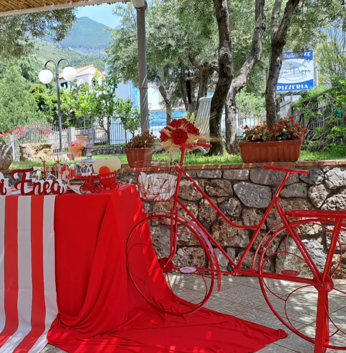 Decorazione con bicicletta rossa accanto a un tavolo con drappeggio rosso in un caffè con giardino, fiori e alberi.