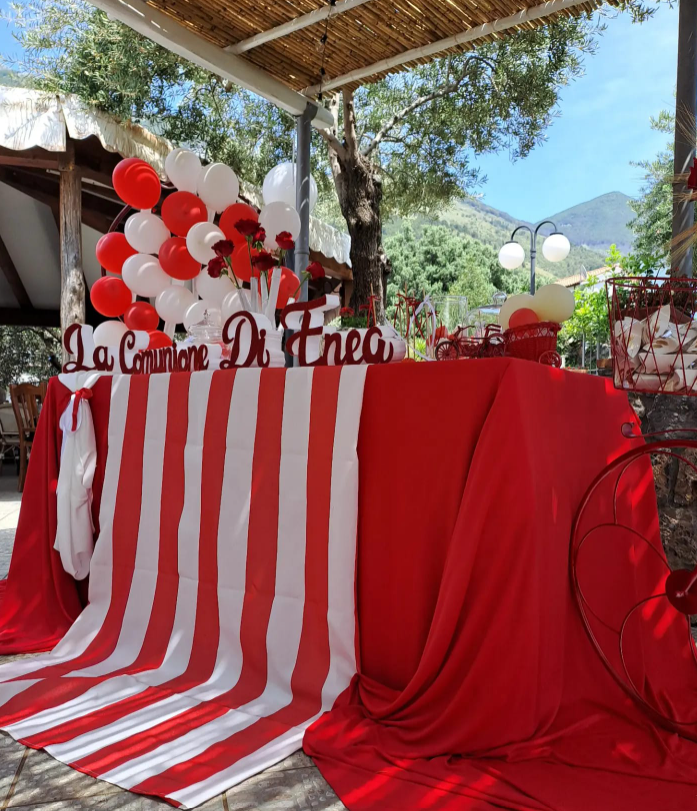 Tavolo da festa decorato in rosso e bianco con palloncini e cartello con il nome, in un ambiente esterno.