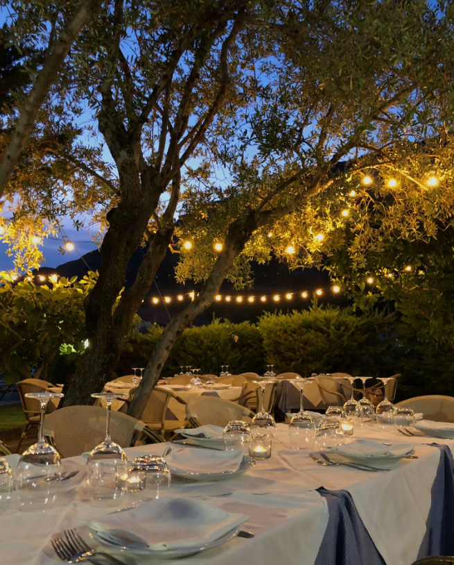Tavolo da pranzo all'aperto apparecchiato sotto luci decorative e alberi al crepuscolo