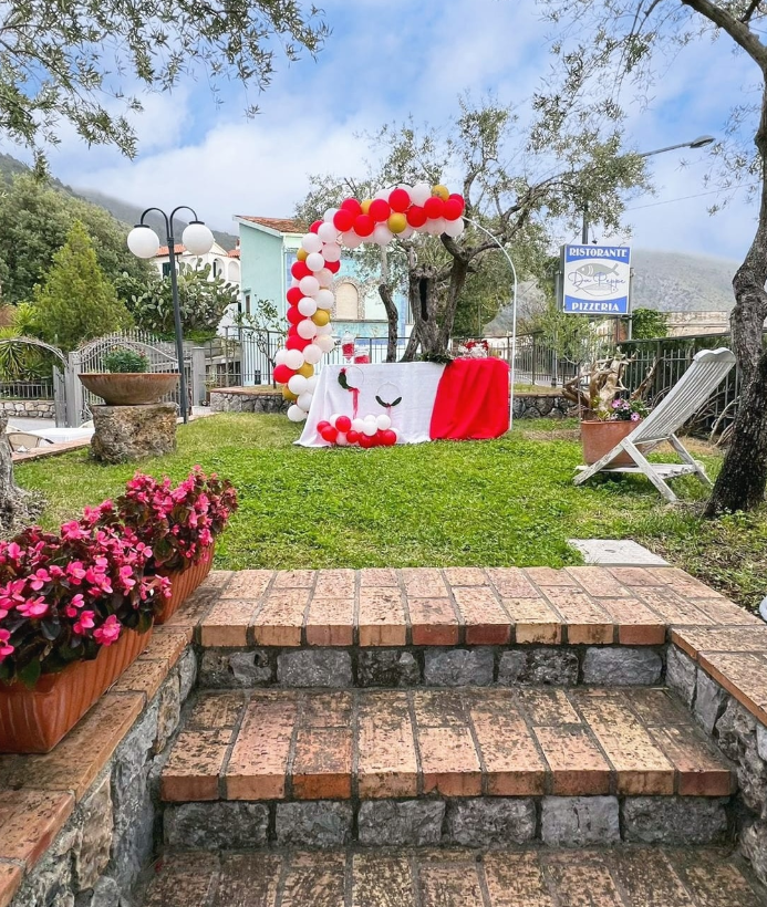 Allestimento da giardino all'aperto con arco di palloncini rossi e bianchi, tavolo decorato e sedie bianche su un prato.