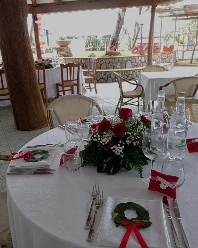 Apparecchiare la tavola con tovaglie bianche, un centrotavola floreale rosso, bicchieri e sedie da patio in riva all'acqua.