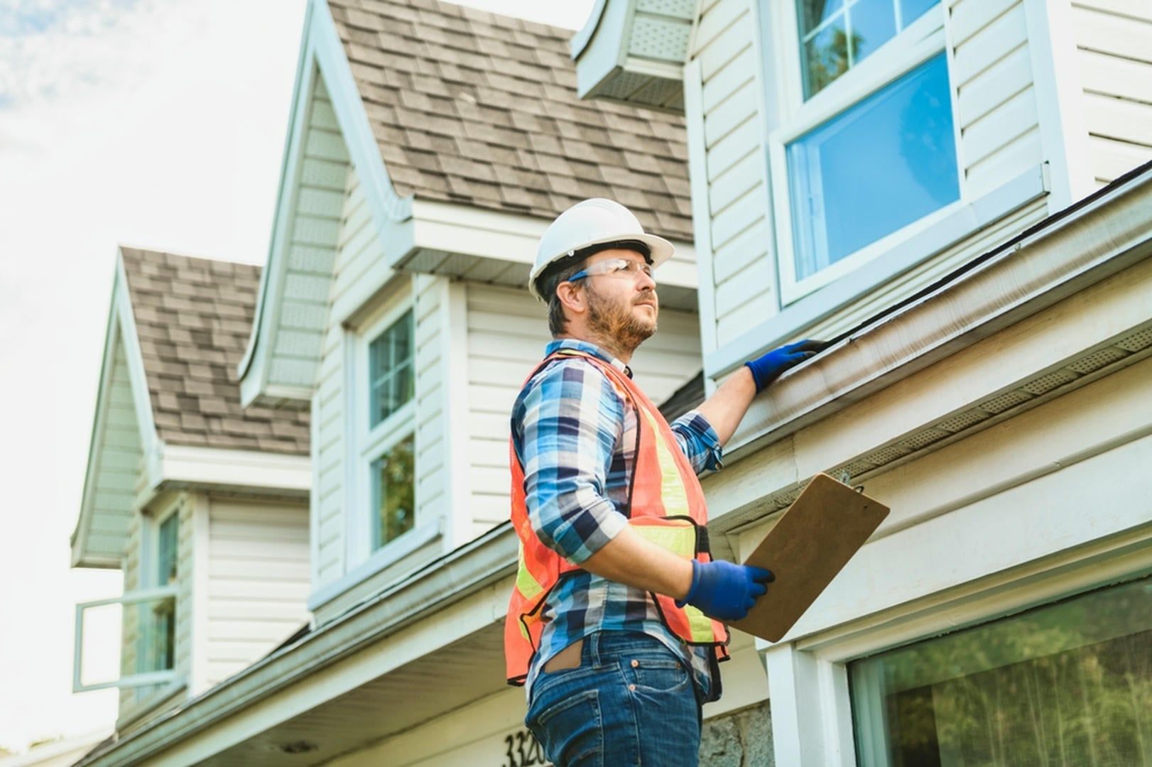 Construction worker inspecting a house exterior, wearing safety gear and holding a clipboard.