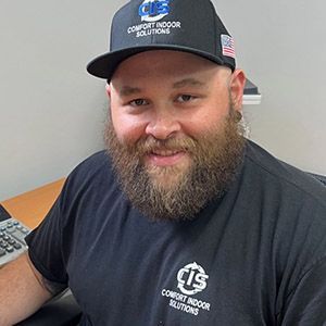 Smiling bearded man in a black cap and T-shirt, seated indoors at a desk with computer equipment.