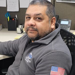 Man seated at an office desk wearing a gray sweater with an American flag patch, looking at the camera.