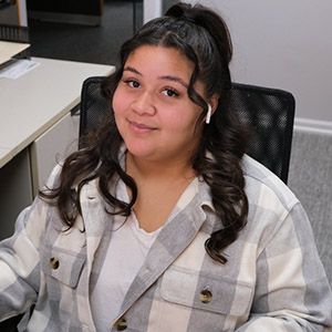 Person seated at a desk, smiling in a gray plaid jacket with dark curly hair.
