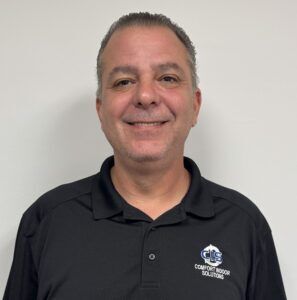 Smiling man in a black polo shirt with a logo, standing against a plain white wall.
