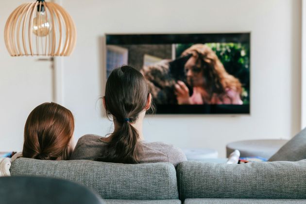 Two people sit on a gray couch watching TV in a bright living room.