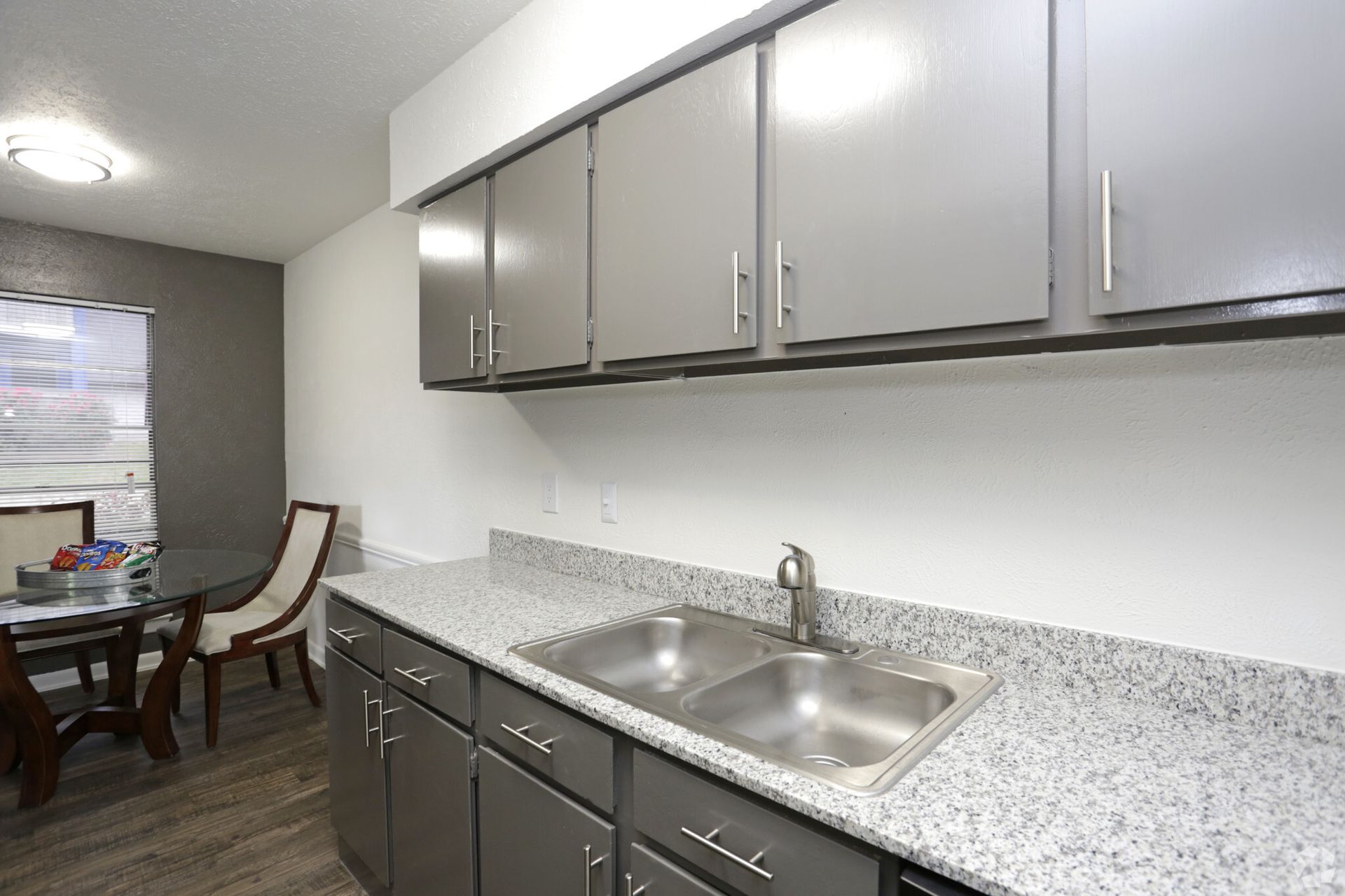 Modern kitchen with gray cabinets, speckled countertops, and stainless steel sink beside a dining area.