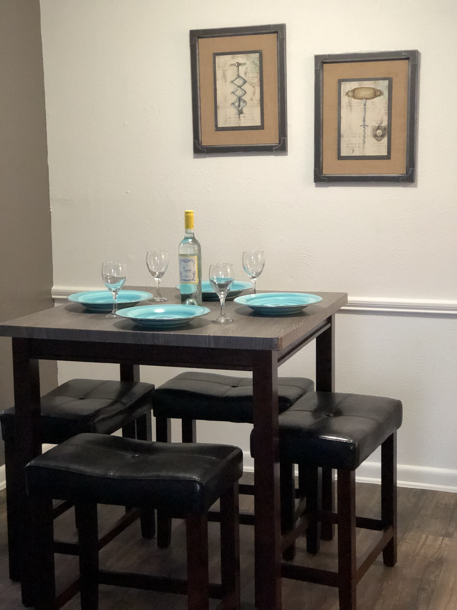 Dining table set for four with turquoise placemats, wine glasses, and wall art in a simple room.