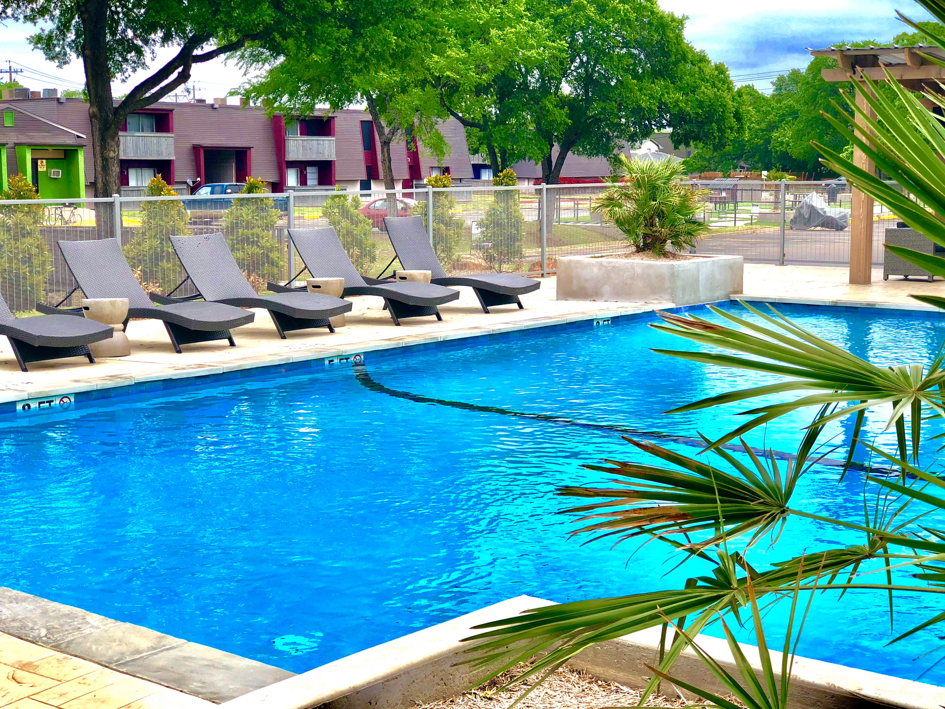 Poolside lounge chairs beside a bright blue swimming pool with trees and a resort building in the background