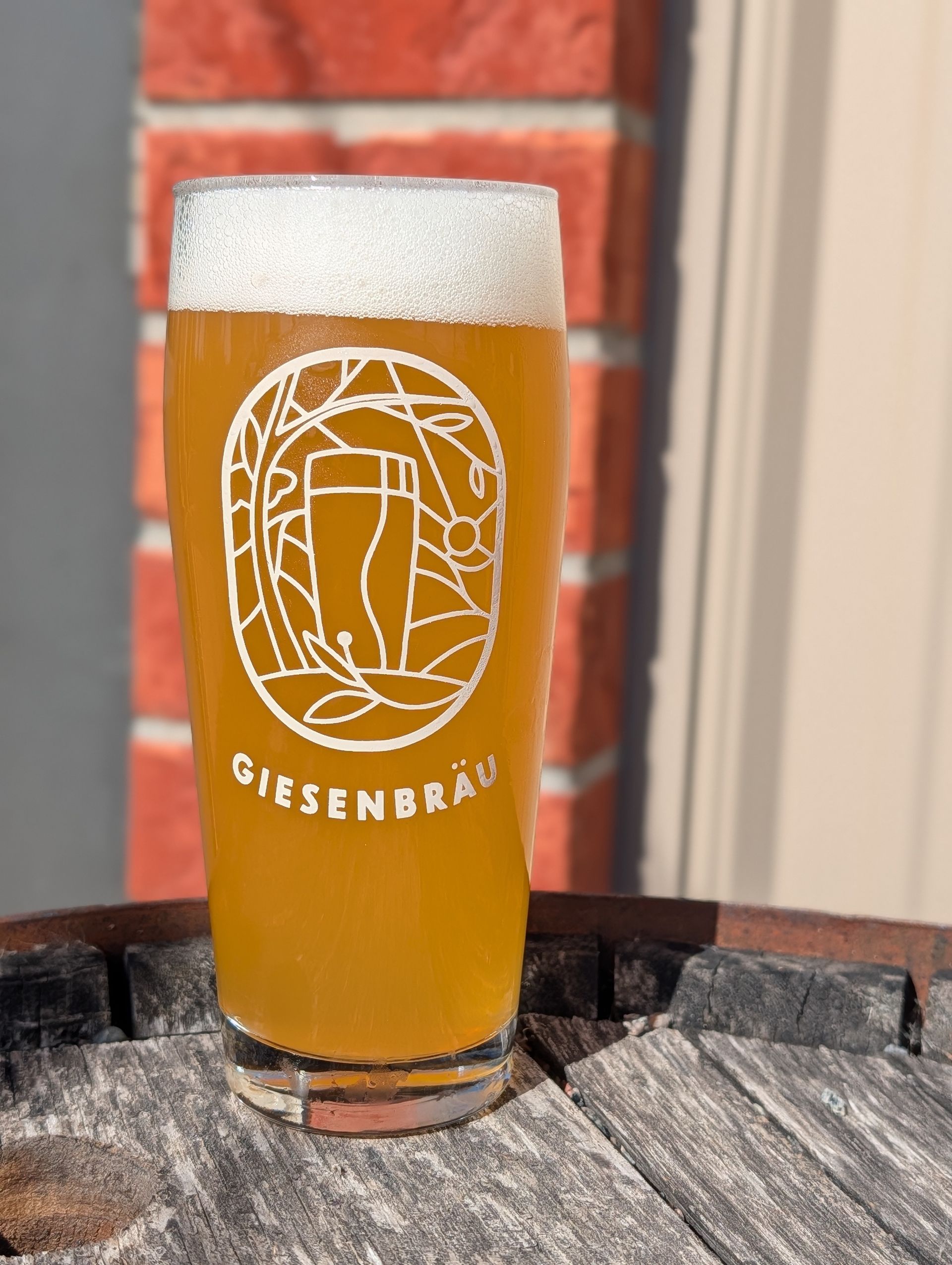 Tall beer glass with a frothy head and Giesenbräu logo on a wooden barrel, against a brick wall