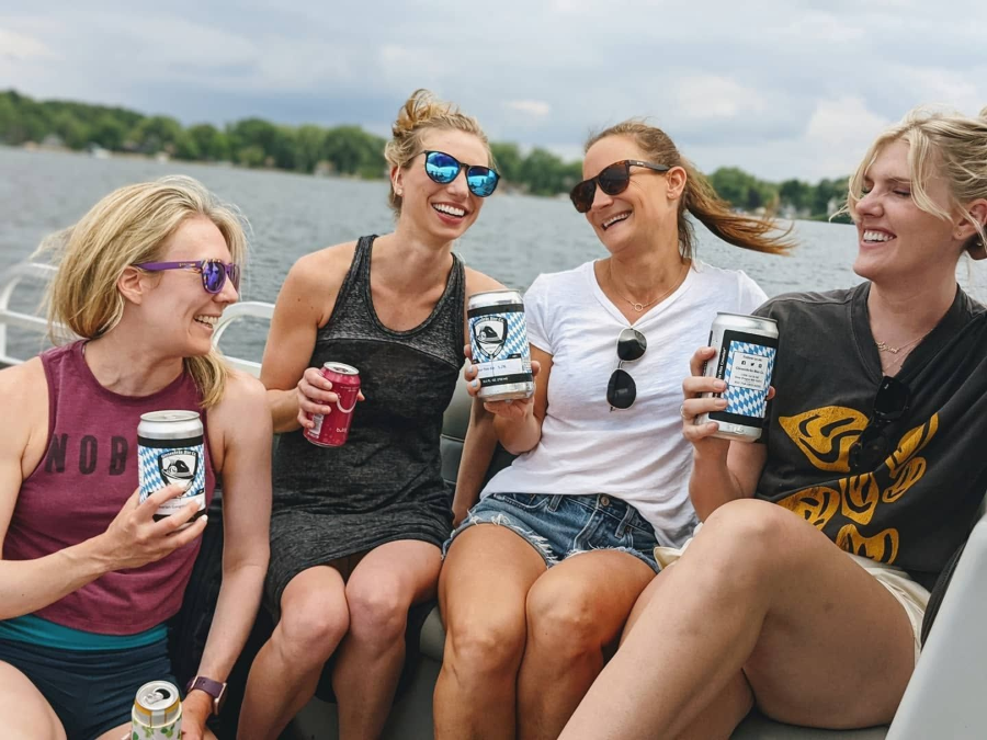 Four friends sit on a boat, smiling and holding drinks on a sunny lake.