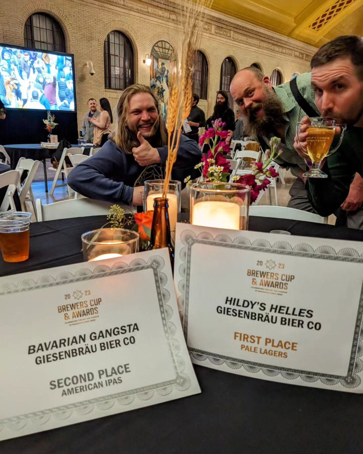 Two men at a dinner table with award placards and drinks in a warmly lit restaurant
