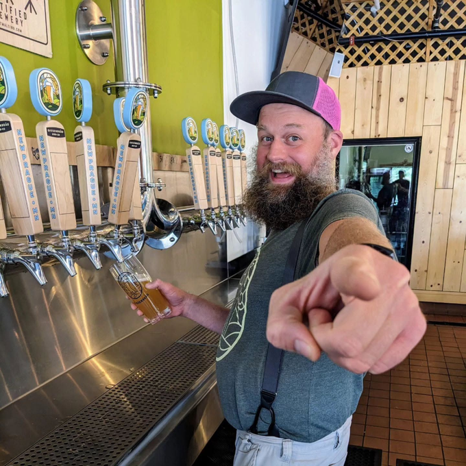 Man at a bar taps a beer and points at the camera, smiling in a brewery taproom.