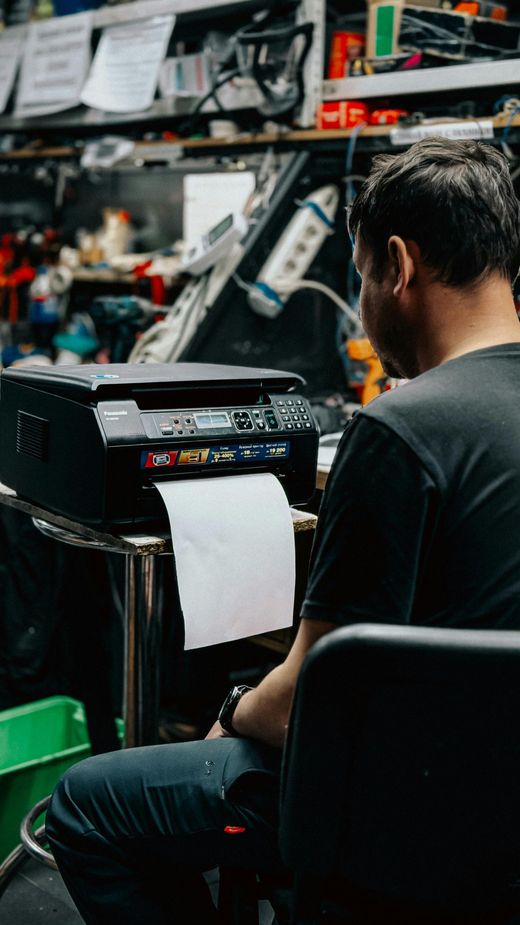 Person working on a printer in a cluttered repair shop, with tools and equipment in the background.