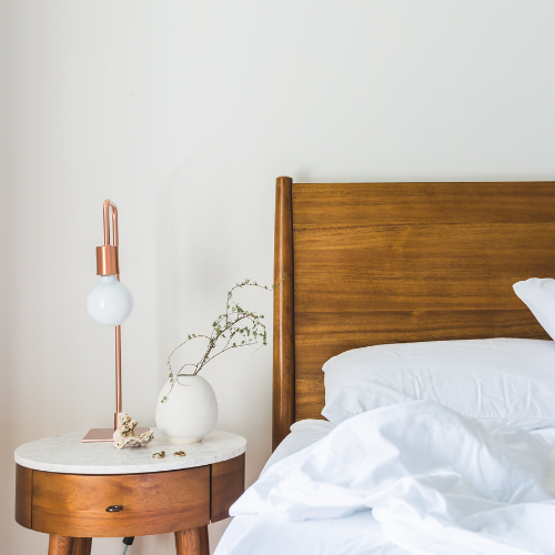Wooden bed with white bedding, round nightstand with lamp, vase, and decorative items.