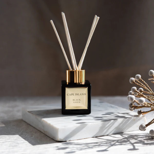 Black glass reed diffuser with gold accents and reeds on a marble square, with a sprig of berries.