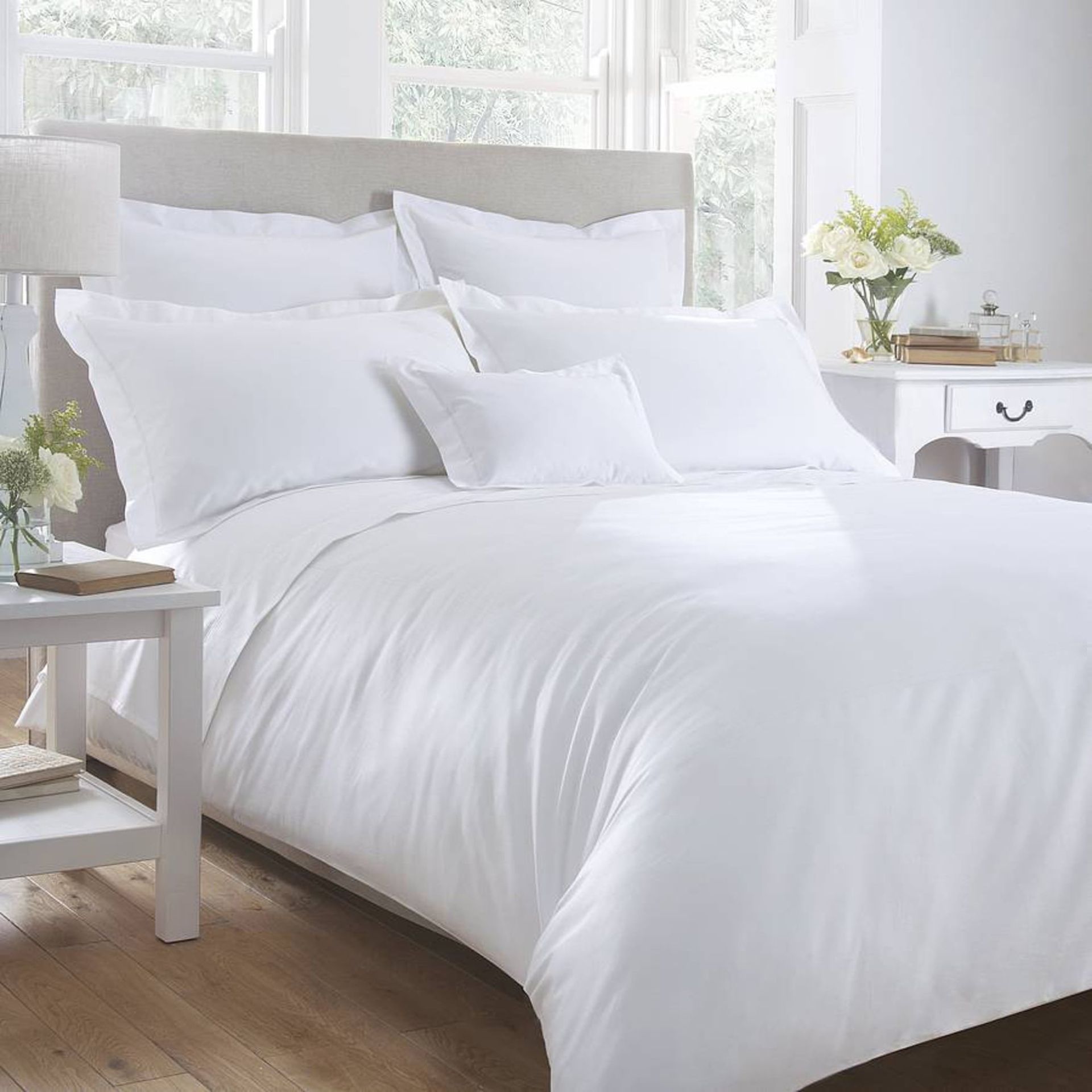 White bed linens in a bright bedroom with a bedside table, lamp, and flowers.