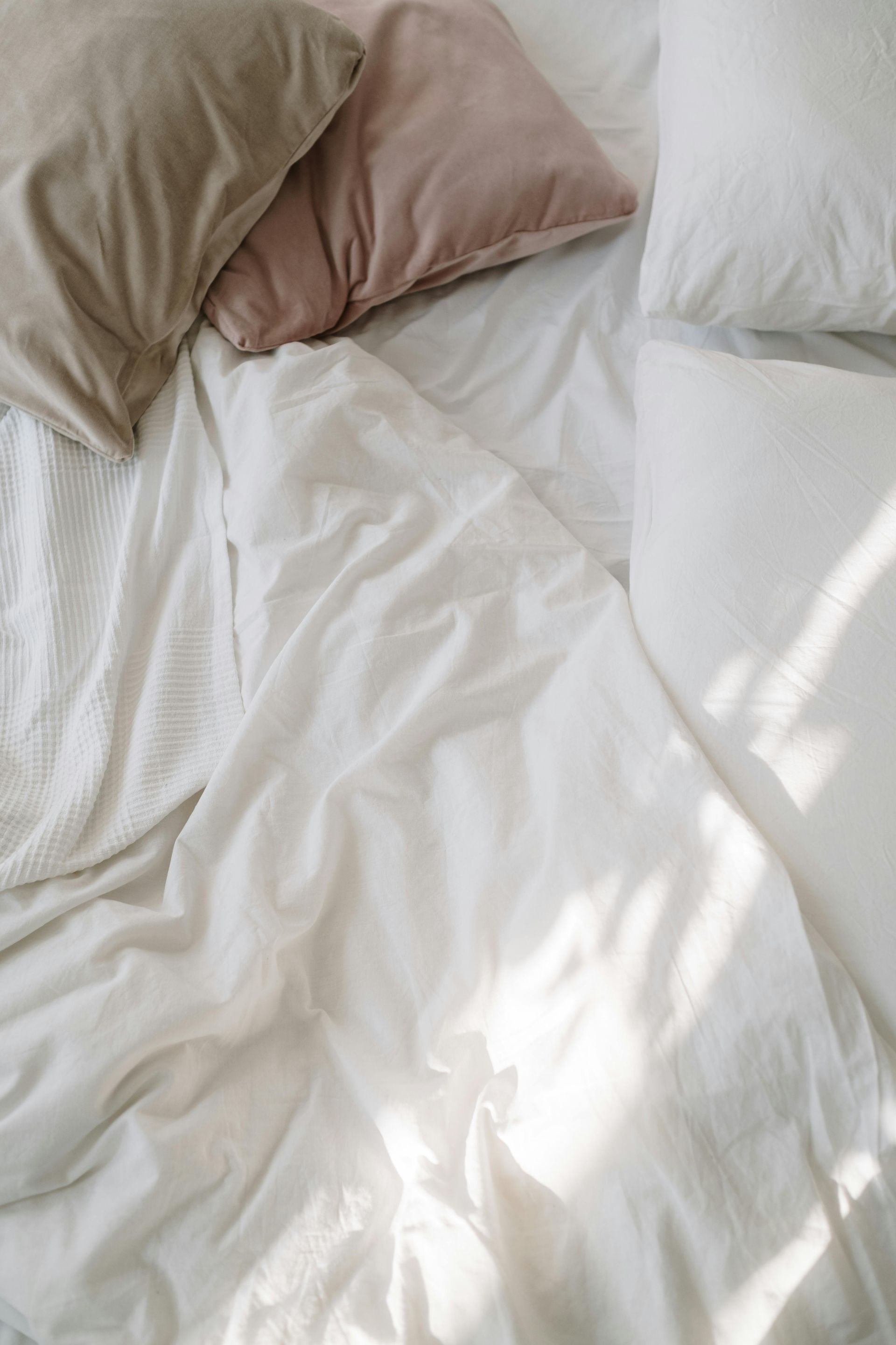 Bed with rumpled white sheets and beige and pink pillows. Sunlight shines.